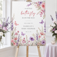 Editable butterfly baby shower welcome sign Canva template with watercolour pink and purple butterflies, peonies, lavender, and wildflower meadow on a white background, shown as a hanging print mockup