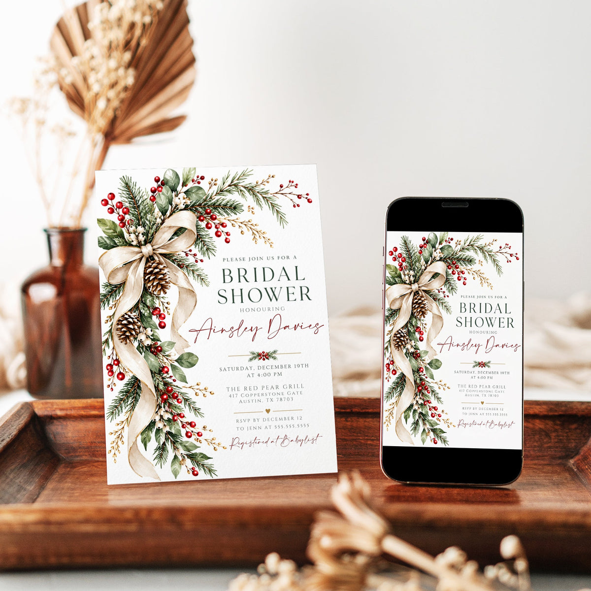 Winter bridal shower invitation with a flowing ivory bow, evergreen branches, red and white berries, snow-dusted pinecones, and gold accents on a white background