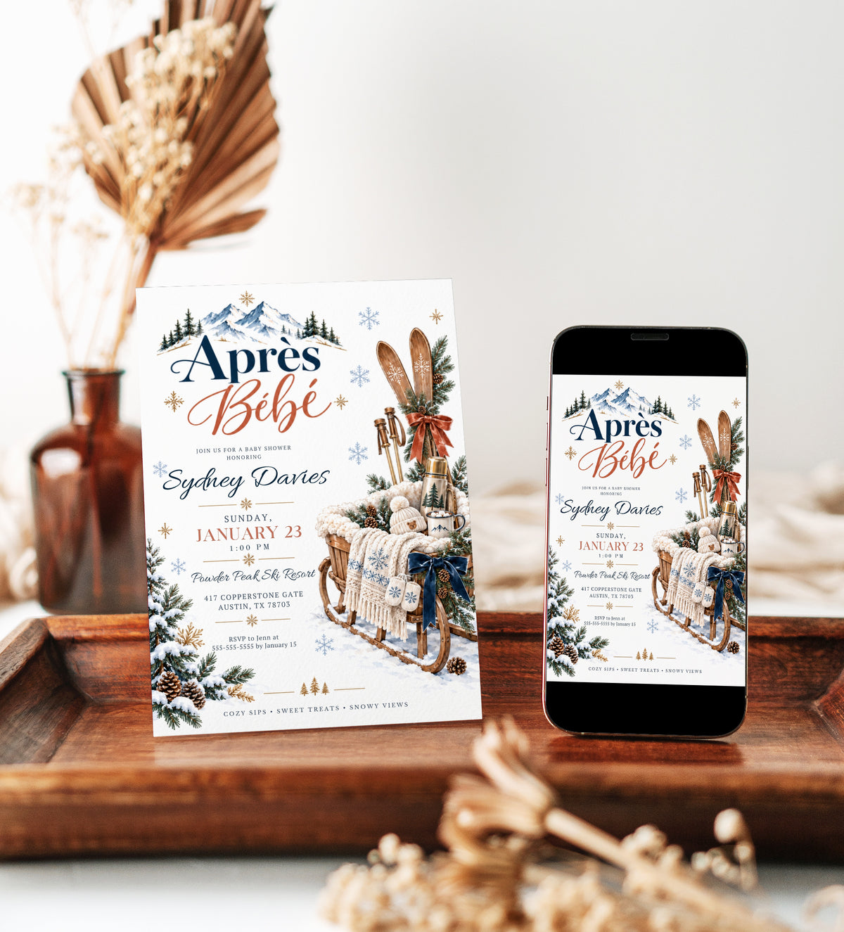 Après Bébé baby shower invitation with wooden skis, a rustic sled, knit baby hat and mittens, hot cocoa, snowy mountains, and evergreen branches