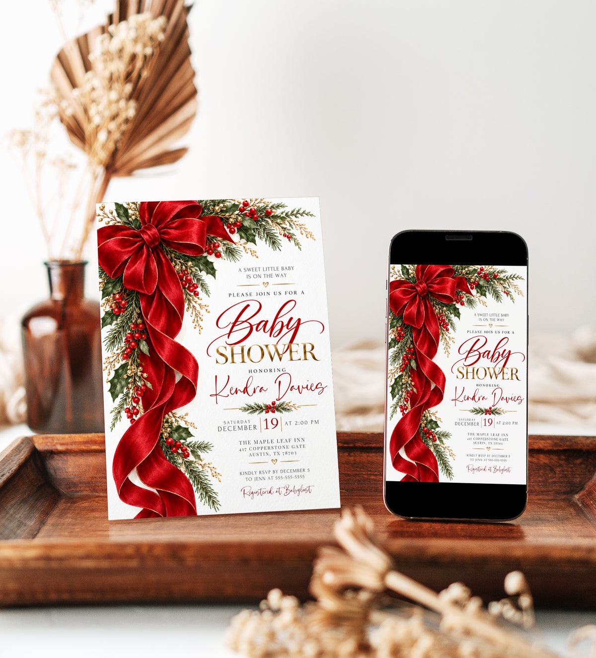 Christmas baby shower invitation with a large red bow and flowing ribbon, green holly, red berries, evergreen branches, and gold lettering on a white background