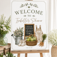Homegrown baby shower welcome sign with a watering can, green apples, and baby overalls in sage green and neutral