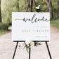 Minimalist black and white horizontal wedding welcome sign template with bold calligraphy script and spaced typography, editable in Canva