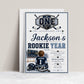 Rookie Year first birthday invitation with little hockey player, ice rink, scoreboard, helmet, hockey sticks, puck, and navy blue birthday party details
