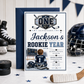 Rookie Year first birthday invitation with little hockey player, ice rink, scoreboard, helmet, hockey sticks, puck, and navy blue birthday party details