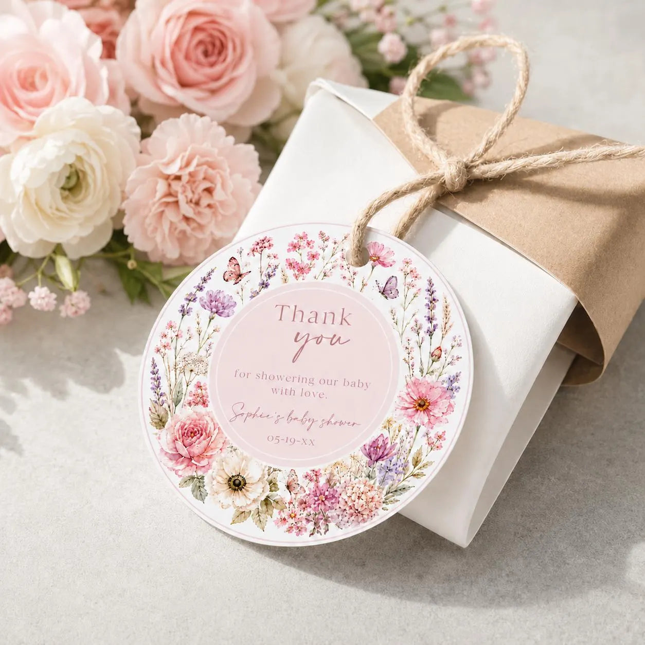 Editable wildflower baby shower round thank you sticker Canva template with watercolour roses, lavender, cosmos, butterfly details, and script typography on a soft blush background, shown tied to a kraft gift box